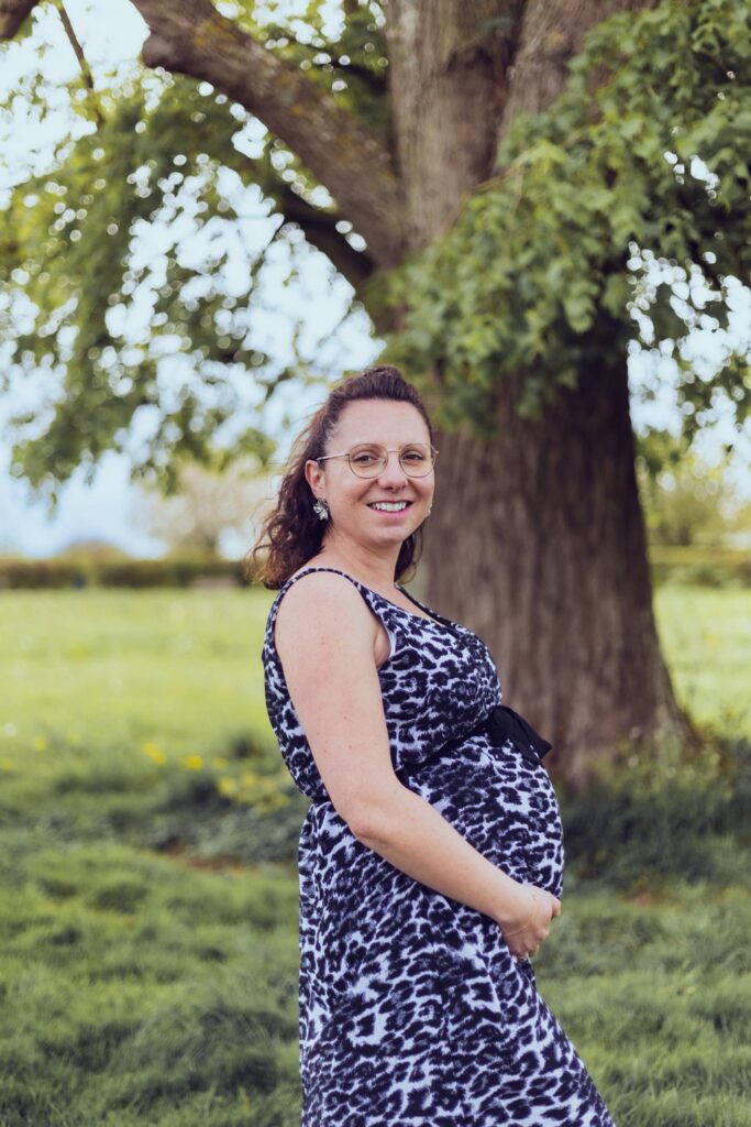 Femme enceinte souriante debout devant un arbre, tenant son ventre dans un parc fleuri.