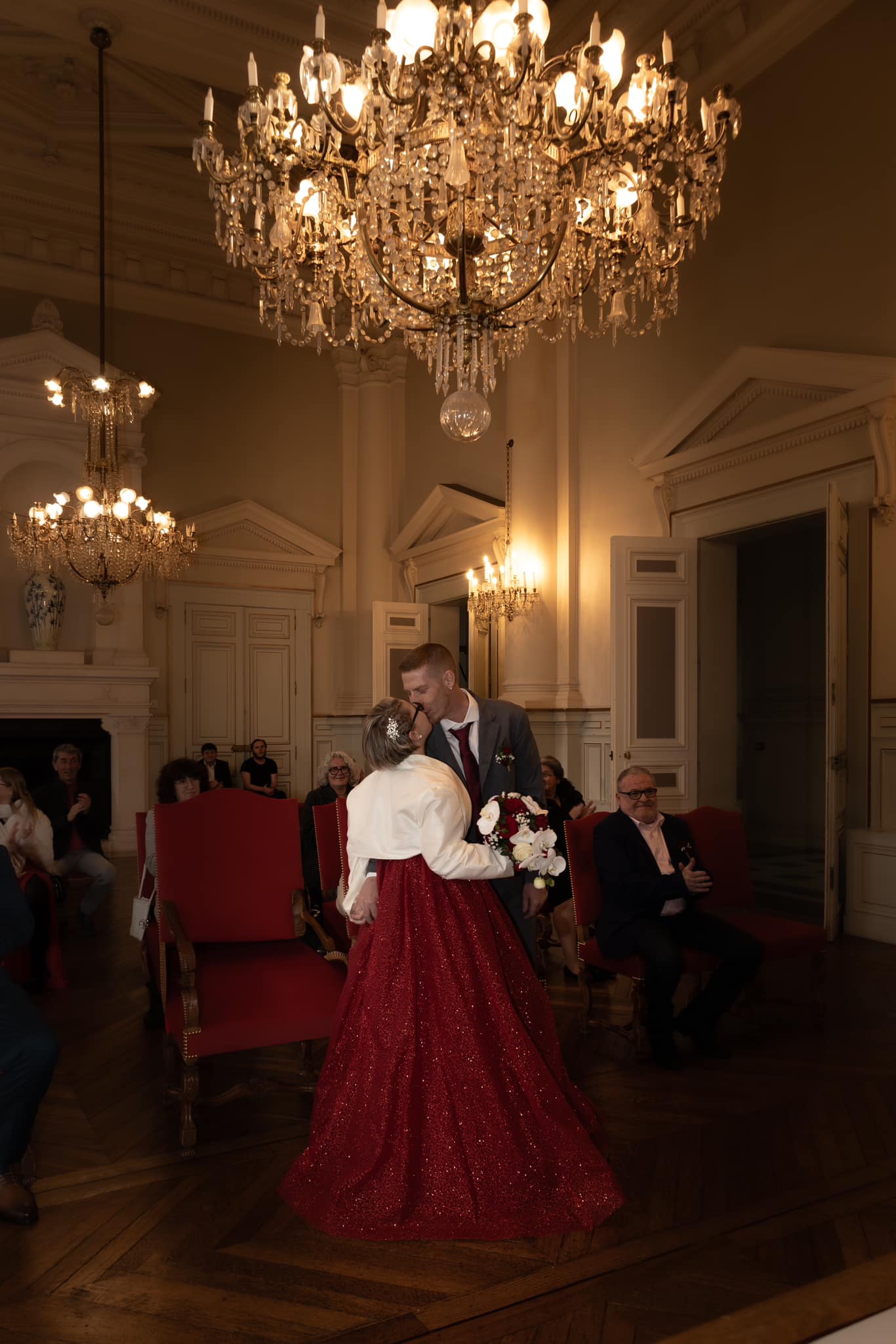 Un couple en tenue de cérémonie s’embrasse dans une salle élégante éclairée par de grands lustres, devant des invités assis.