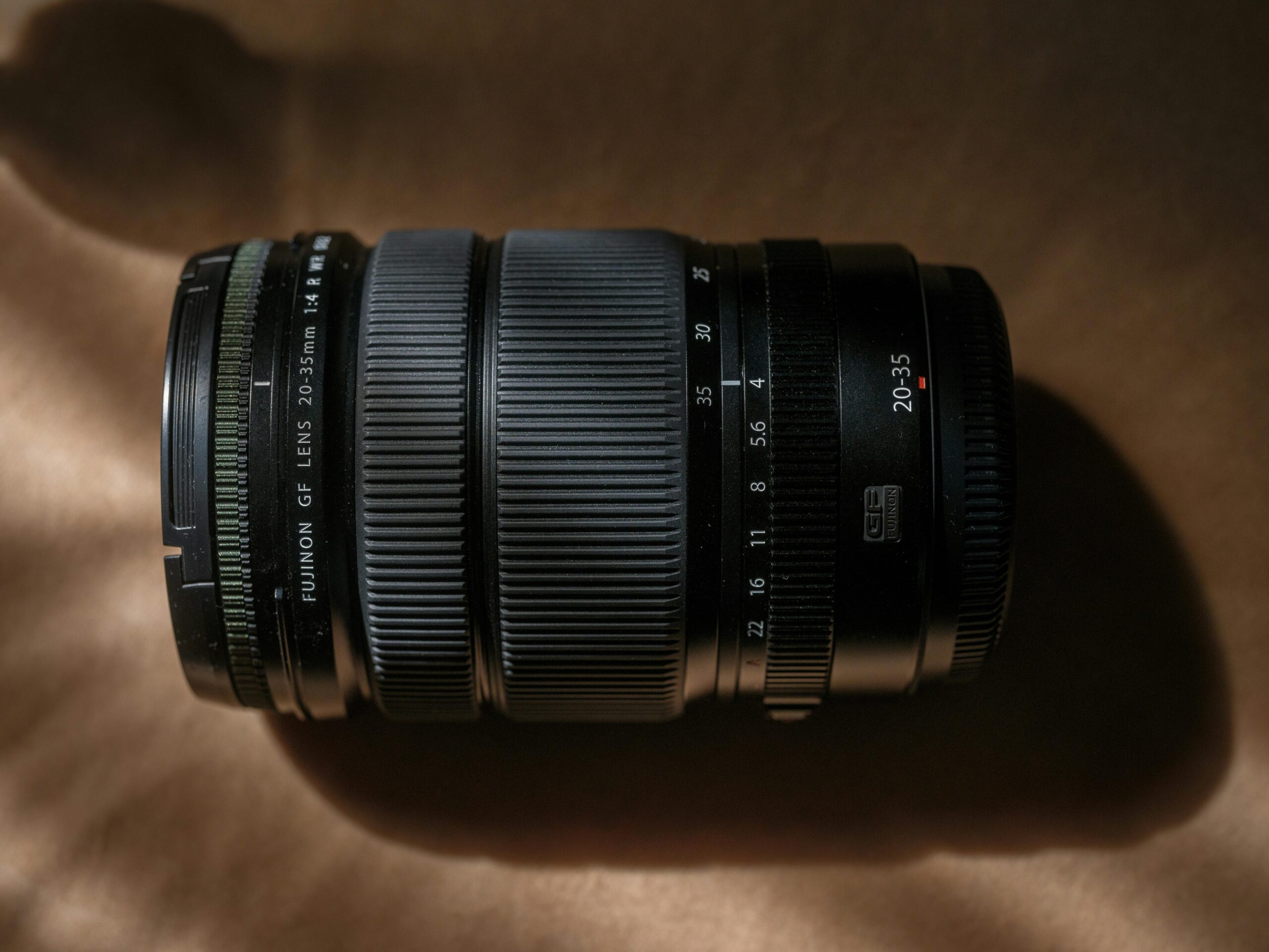 Objectif Fujinon GF 20-35 mm f/4 posé sur une surface brune, éclairé par une lumière dramatique qui souligne ses courbes et ses détails. Les inscriptions sur l’objectif indiquent les focales, les ouvertures et les repères de mise au point.